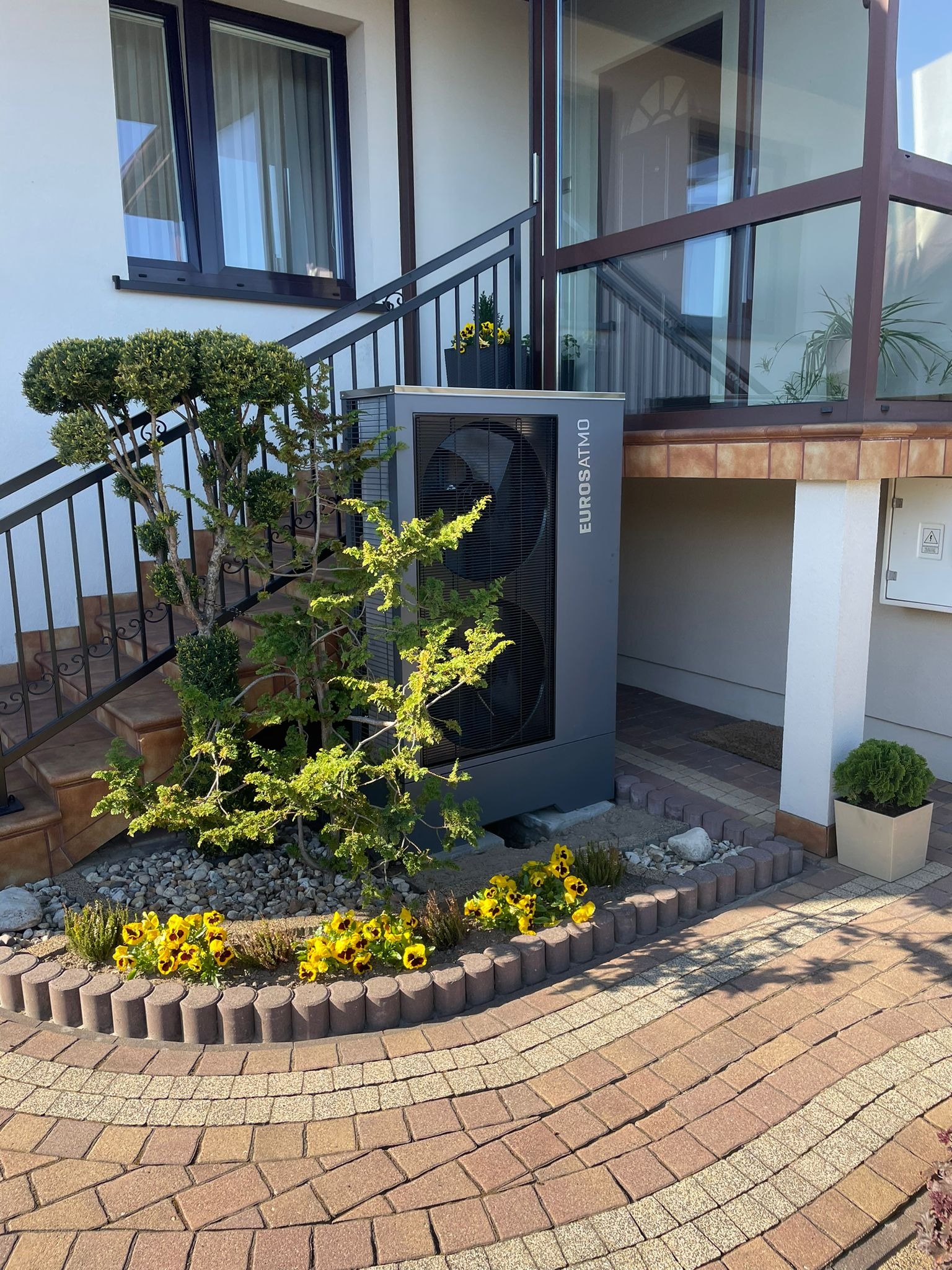 Szara jednostka zewnętrzna z logiem Eurosatmo zainstalowana przy schodach wejściowych do domu w miejscowości Koprki, otoczona ozdobną roślinnością i żółtymi kwiatami.