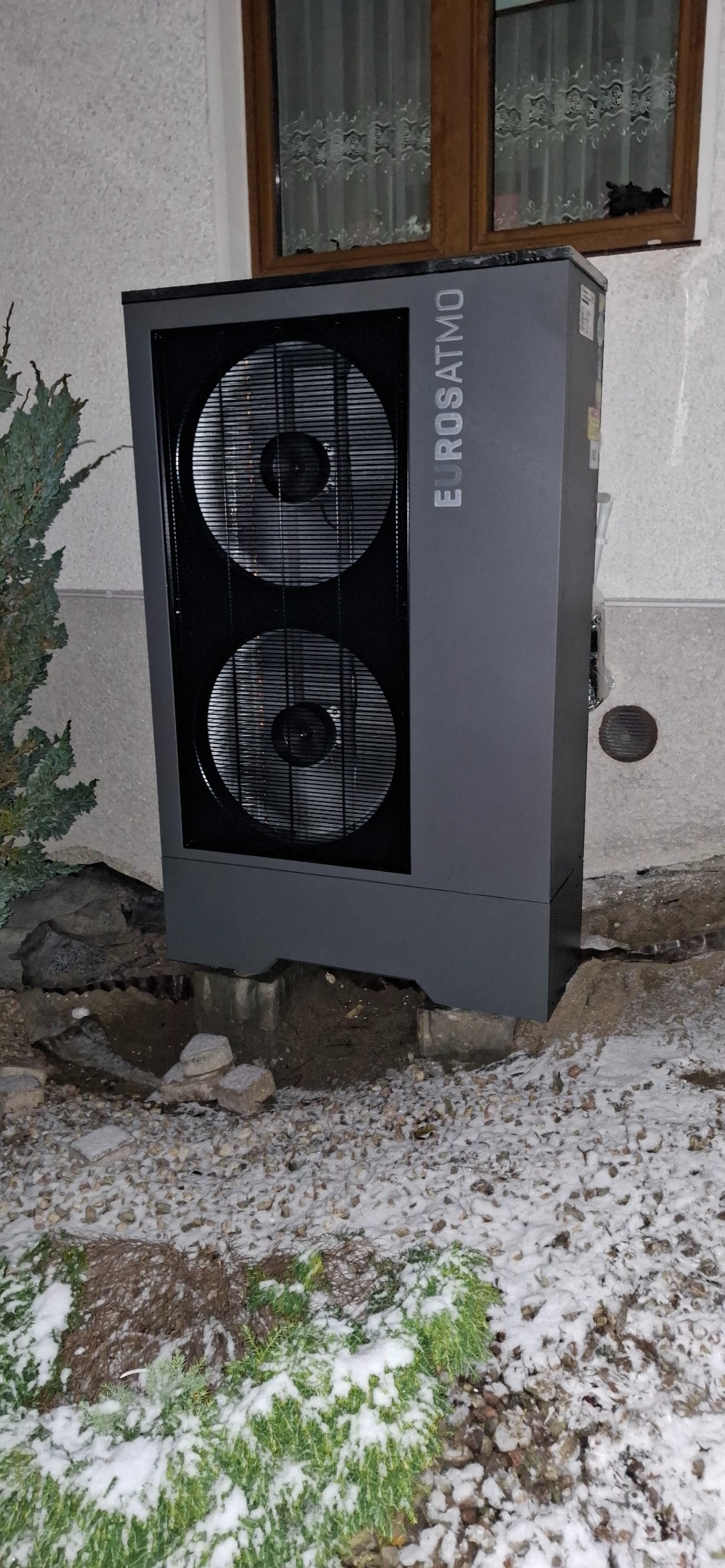 Szara pompa ciepła Eurosatmo z dwoma wentylatorami zainstalowana przy ścianie domu, otoczona kamieniami i zielenią pokrytą śniegiem.
