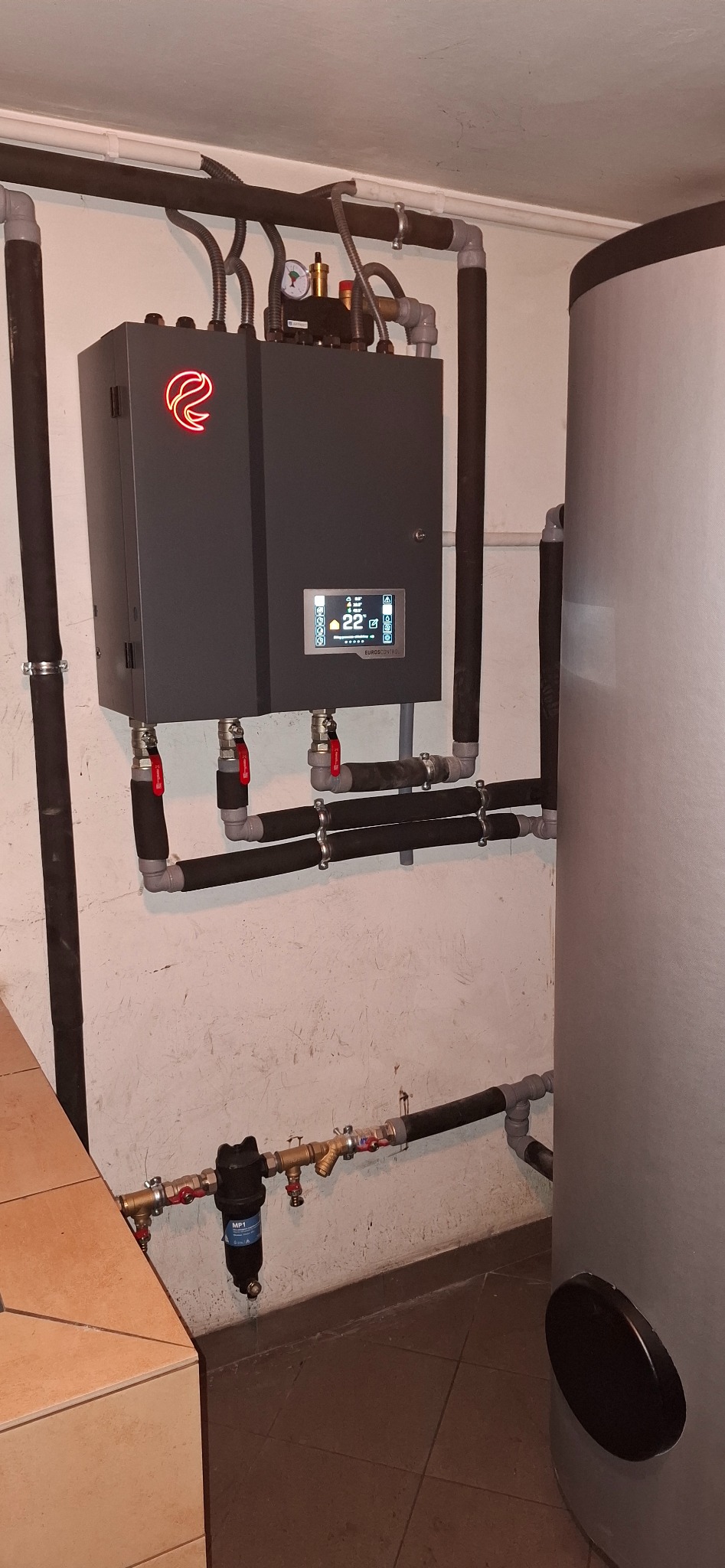 Szara jednostka z podświetlanym czerwonym logo i cyfrowym wyświetlaczem, podłączona rurami do zbiornika akumulacyjnego w piwnicy.
