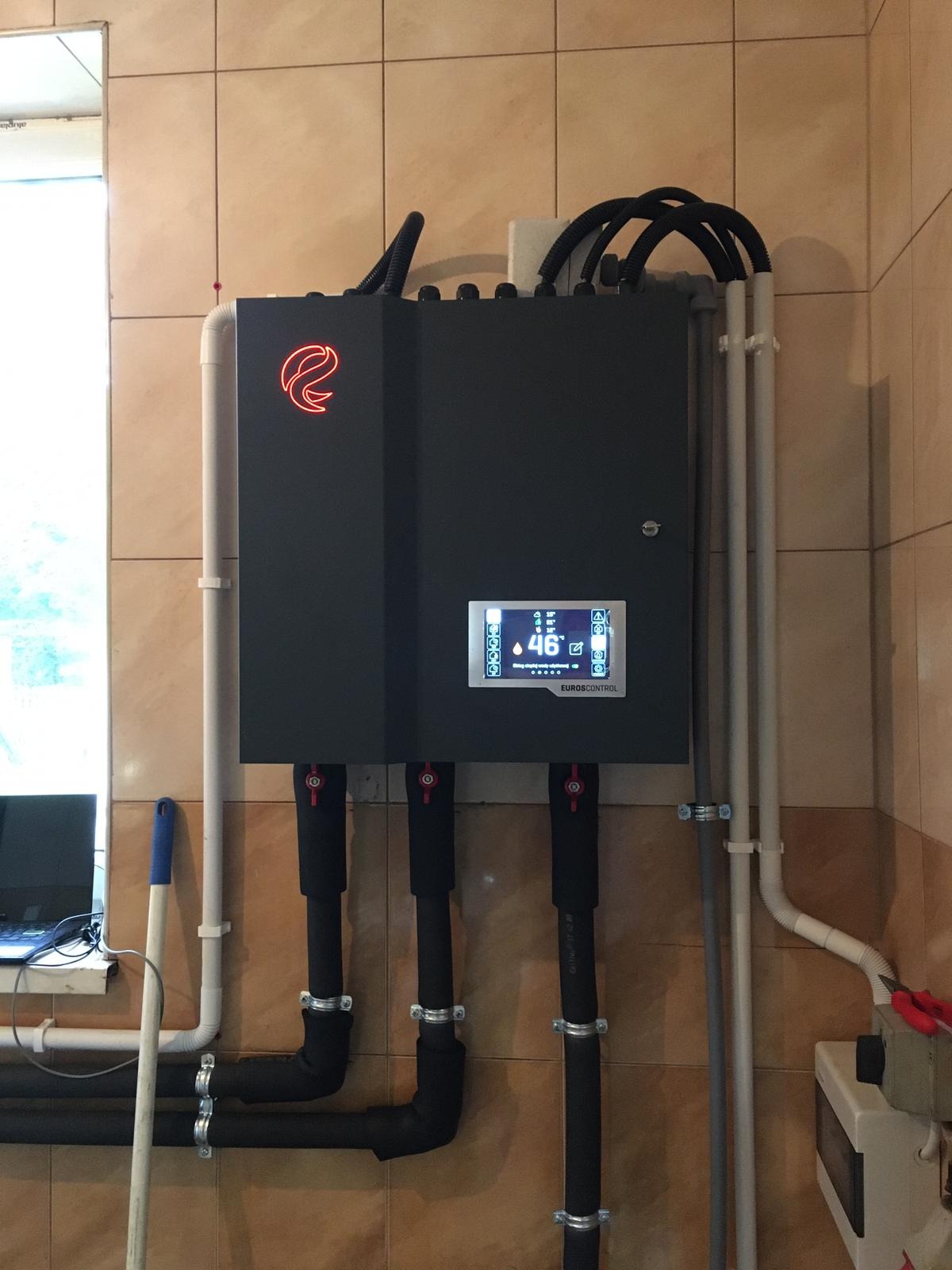 Czarna obudowa urządzenia Eurocontrol z podłączonymi rurami, zamontowana na kafelkowanej ścianie w pomieszczeniu technicznym, widoczny ekran z temperaturą 46 stopni Celsjusza, obok pionowa biała...