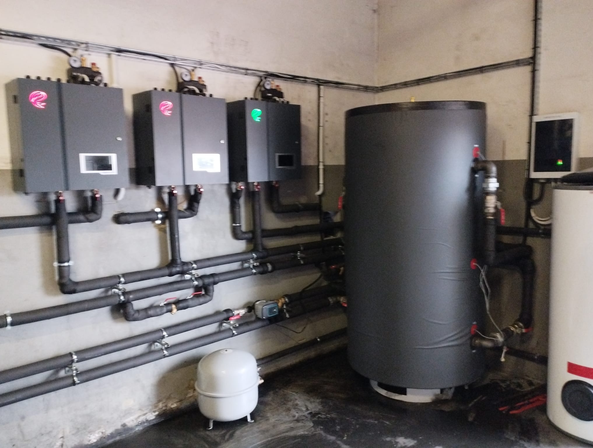 Trzy szare urządzenia z różowymi i zielonymi logotypami na ścianie, połączone czarnymi, izolowanymi rurami, obok duży, czarny zbiornik akumulacyjny i mniejszy, biały zbiornik.