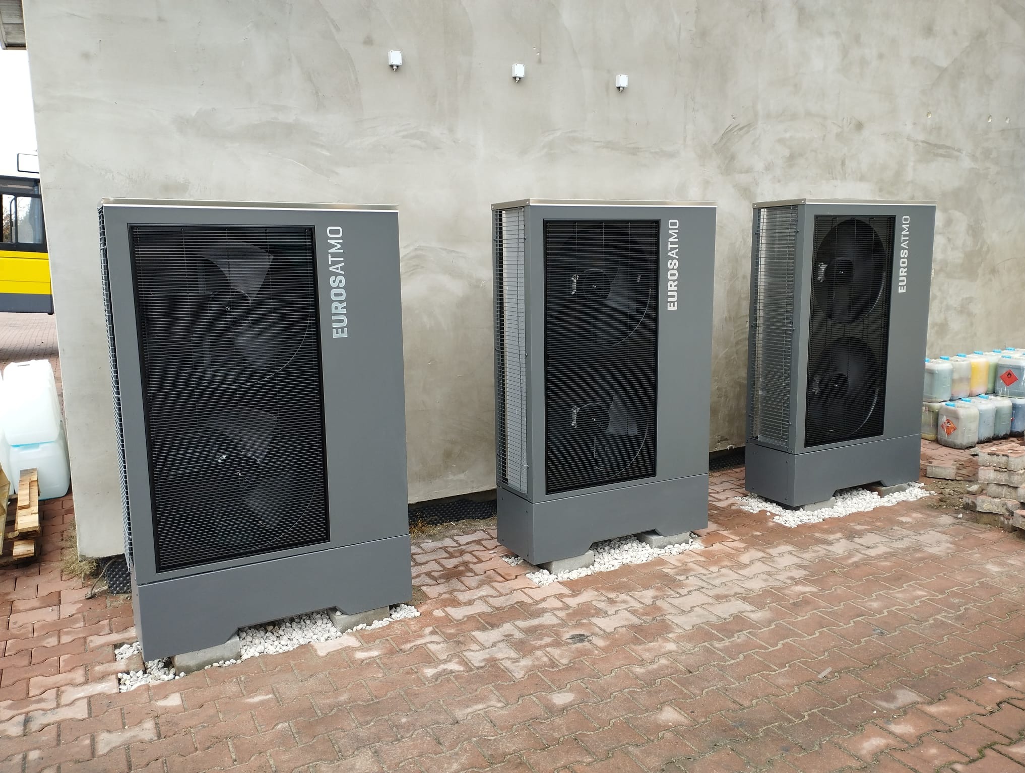 Trzy szare urządzenia grzewcze Eurosatmo ustawione obok siebie na kostce brukowej, z białym żwirem wokół podstawy, na tle szarej ściany. Widoczne pojemniki z płynami w tle.