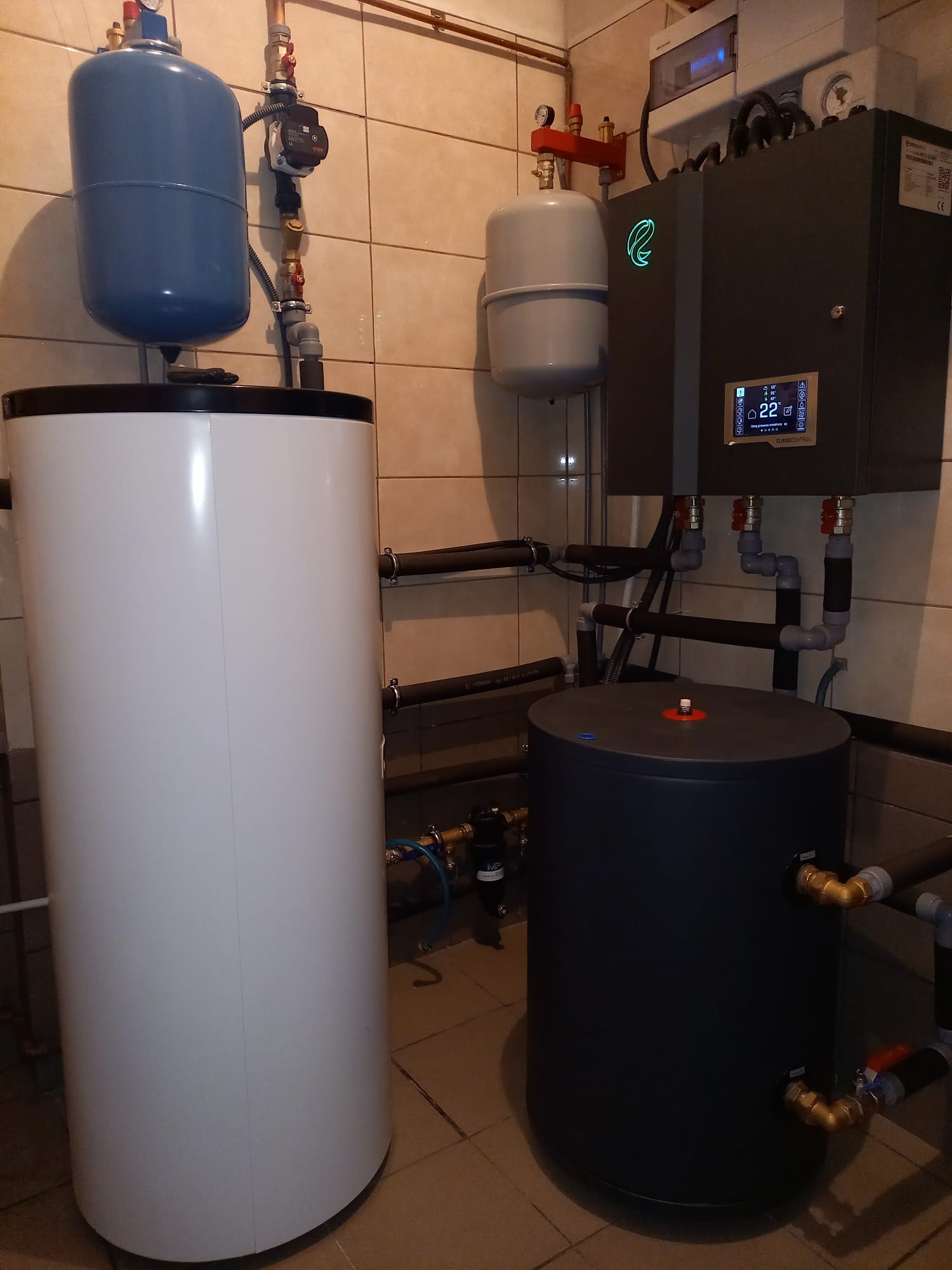 Instalacja w kotłowni z białym zasobnikiem ciepłej wody, szarym zbiornikiem buforowym, szarym modułem sterującym z podświetlanym logo i wyświetlaczem temperatury oraz niebieskim naczyniem wzbiorczym.