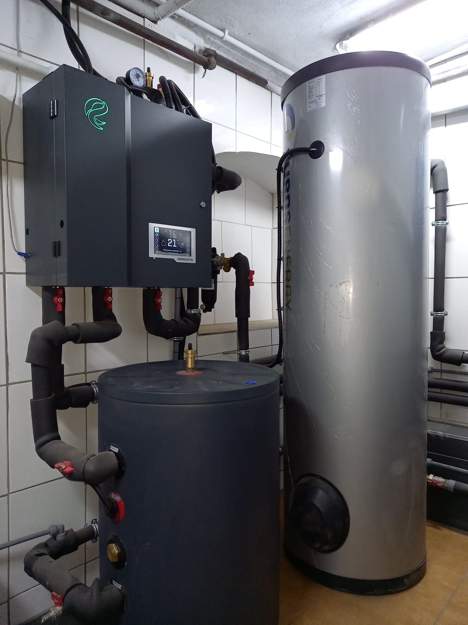Zestaw urządzeń: czarny panel sterowania z zielonym logo, podłączony rurami do szarego i czarnego zbiornika w kotłowni z białymi kafelkami.
