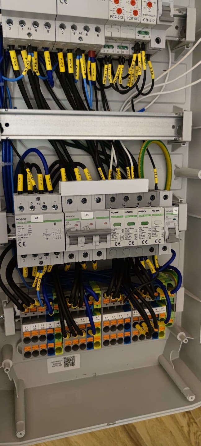Szczegółowe ujęcie rozdzielnicy elektrycznej z widocznymi przewodami oznaczonymi żółtymi etykietami, zabezpieczeniami NOARK i listwami zaciskowymi w kolorach niebieskim, żółtym i pomarańczowym.
