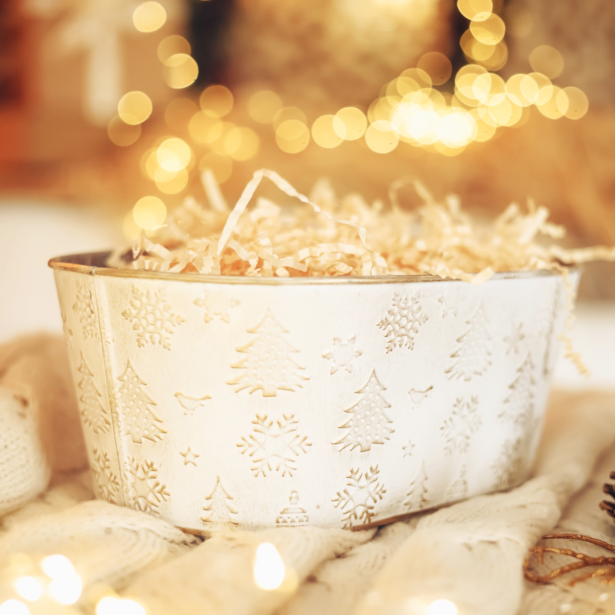 Metalowa, kremowa donica z wytłoczonymi wzorami choinek i płatków śniegu, wypełniona jasnym, papierowym wypełniaczem, umieszczona na puszystym, kremowym pledzie, oświetlona ciepłymi, rozmytymi...