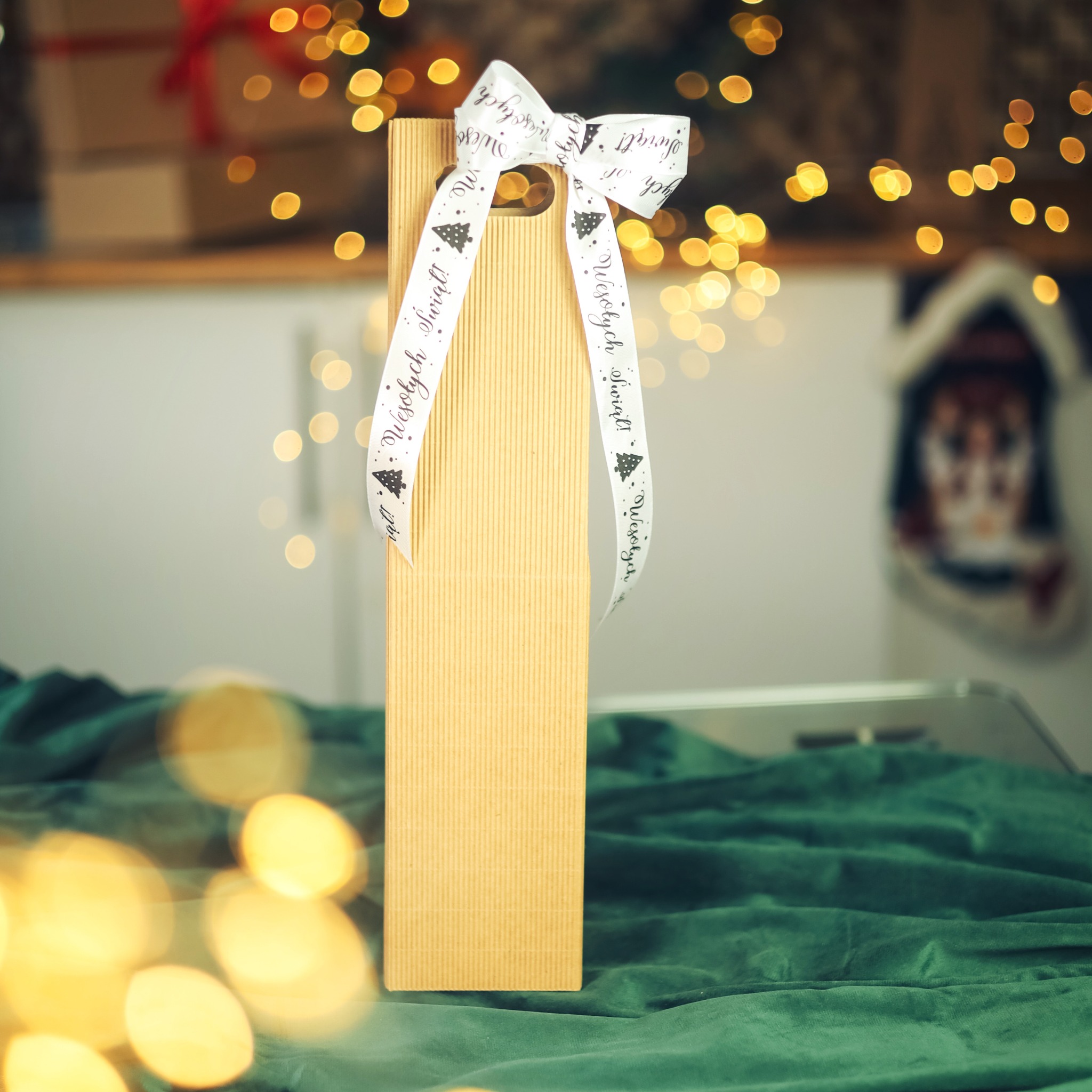 Podłużne pudełko z tektury, przewiązane białą wstążką z napisem 'Wesołych Świąt!' i motywem choinki, na zielonym aksamitnym tle, w tle dekoracje świąteczne i lampki.