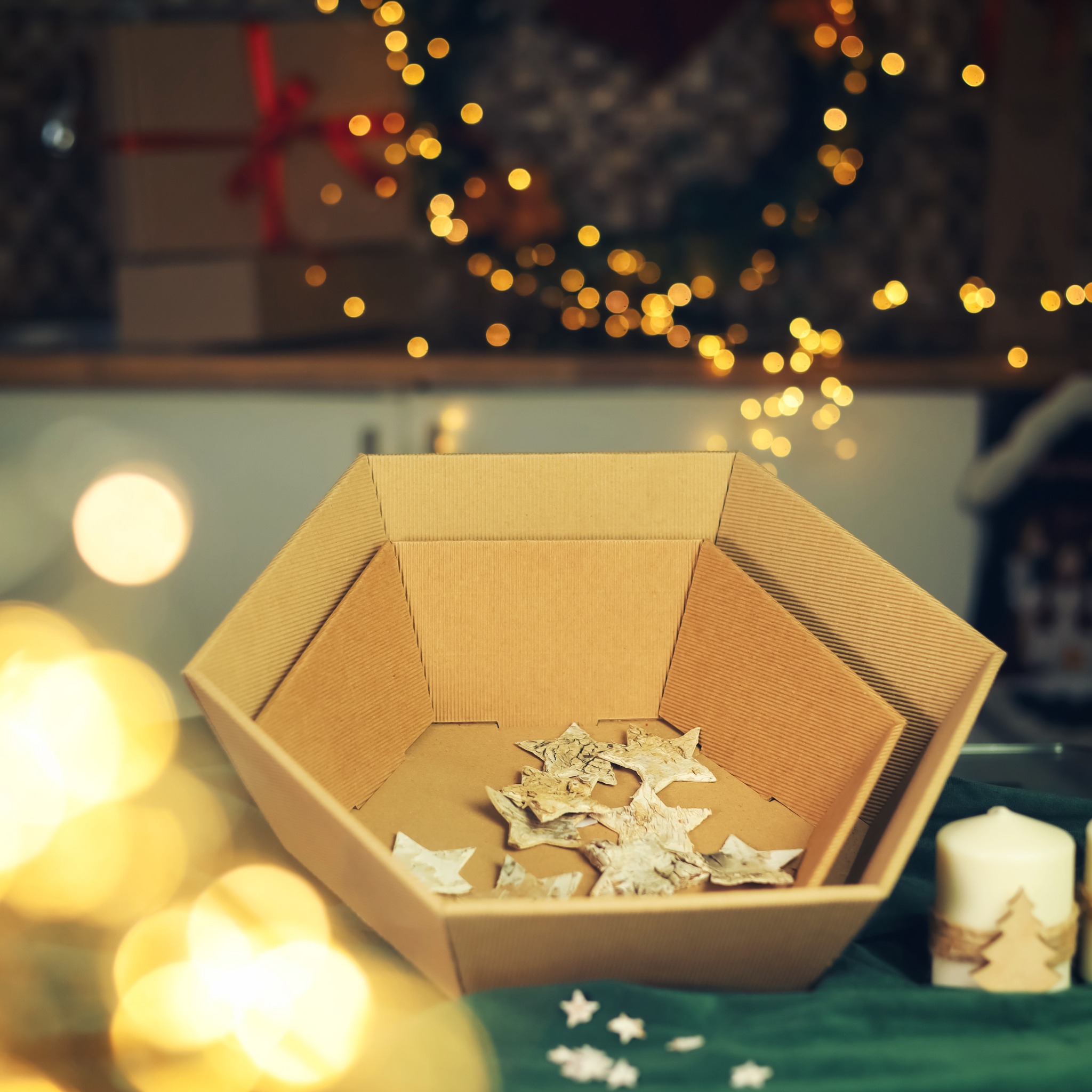 Otwarty, tekturowy pojemnik w kształcie wielokąta wypełniony dekoracyjnymi gwiazdkami, ustawiony na zielonej tkaninie z drobnymi gwiazdkami, w tle rozmyte lampki choinkowe i zapakowane prezenty.