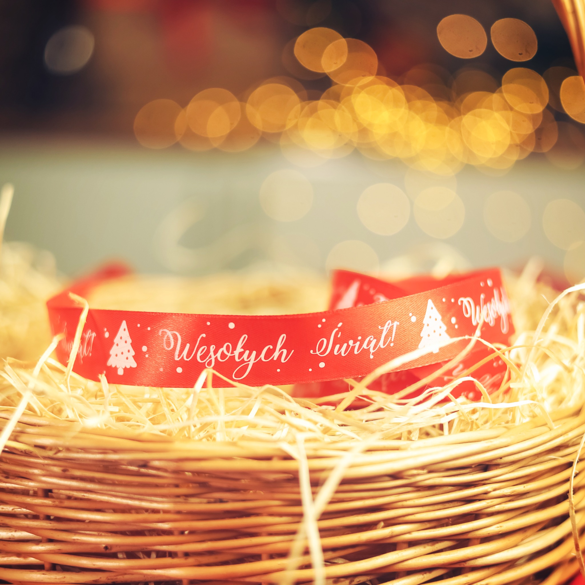 Czerwona wstążka z napisem 'Wesołych Świąt!' i choinkami, leżąca na wypełniaczu w wiklinowym koszu, z rozmytym, ciepłym oświetleniem w tle.