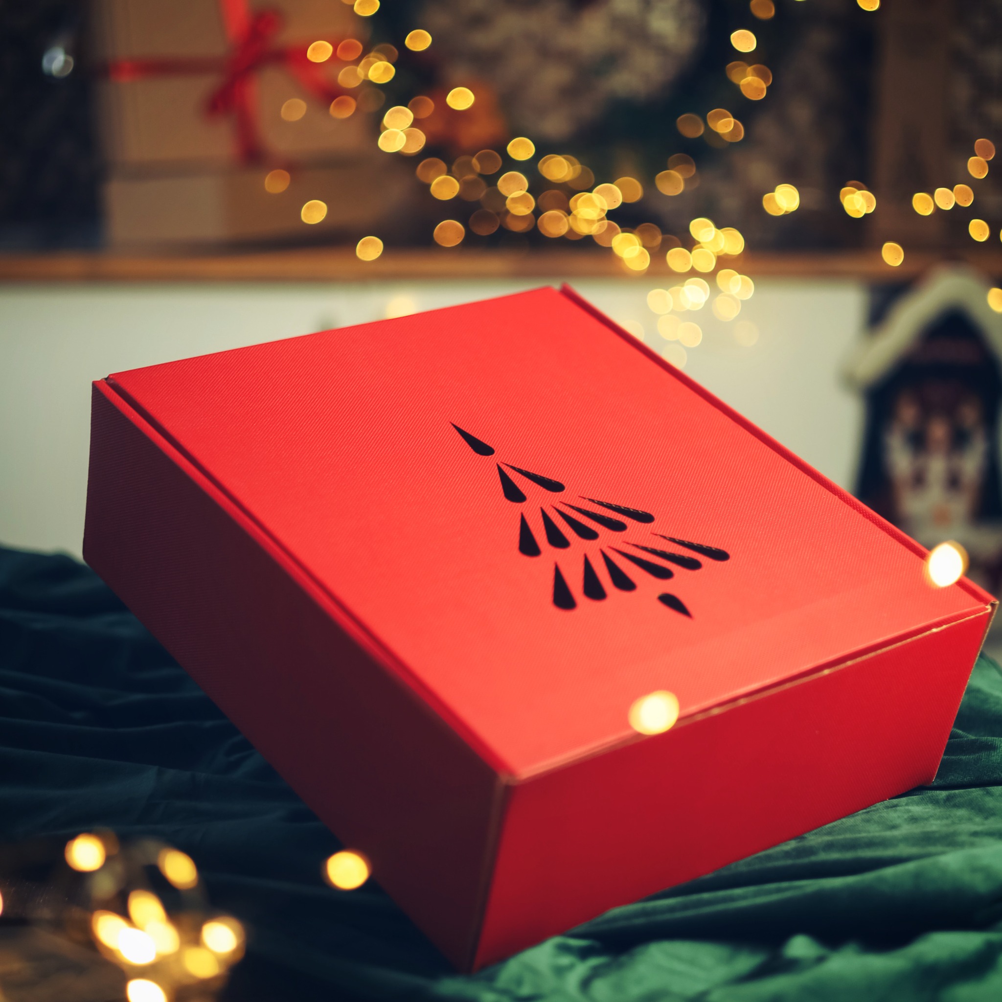Czerwone pudełko prezentowe z motywem choinki na tle zielonego aksamitu i rozmytych światełek.