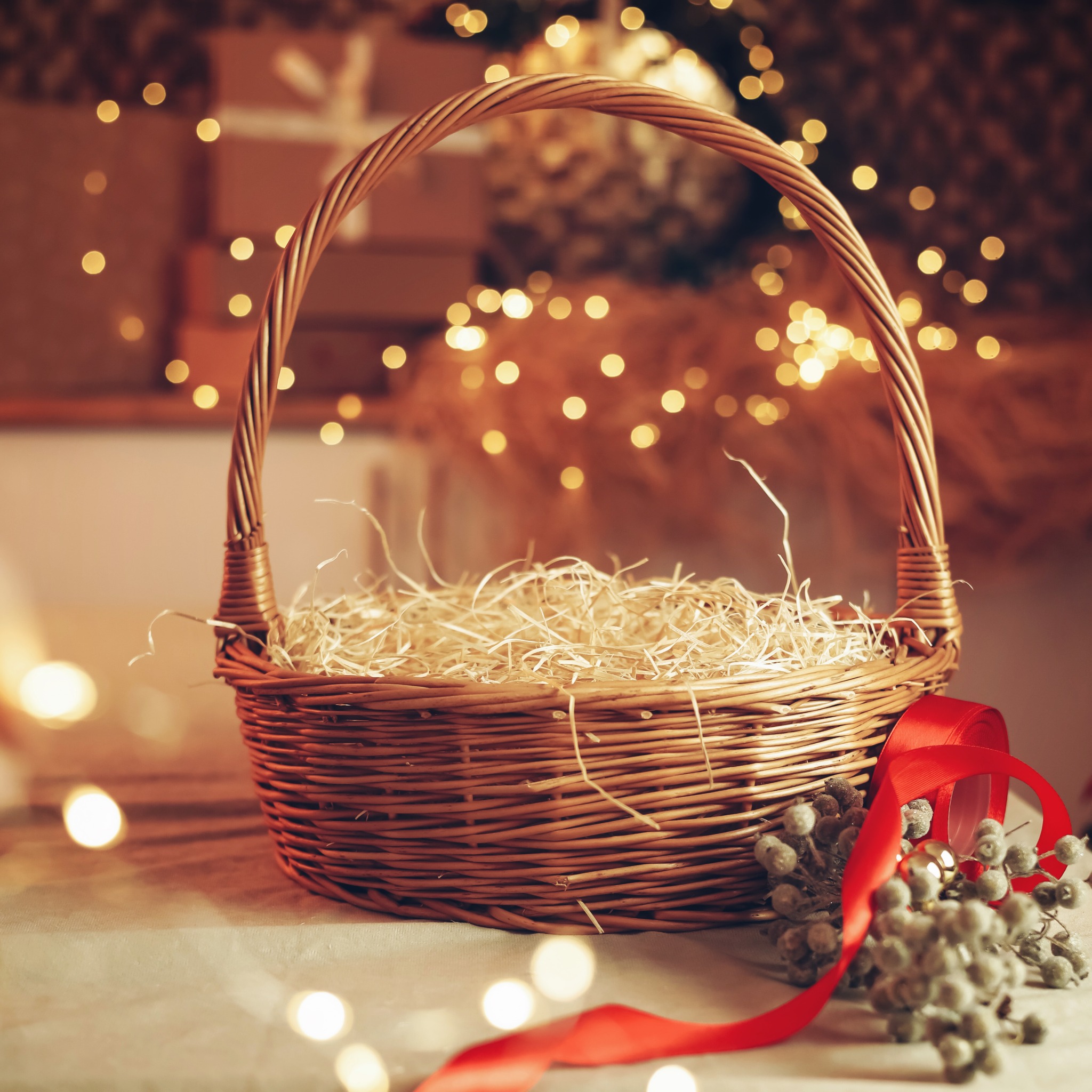Pusty wiklinowy kosz z wypełnieniem z wiórków, ozdobiony czerwoną wstążką i gałązkami z szarymi kuleczkami, na tle rozmytych światełek.