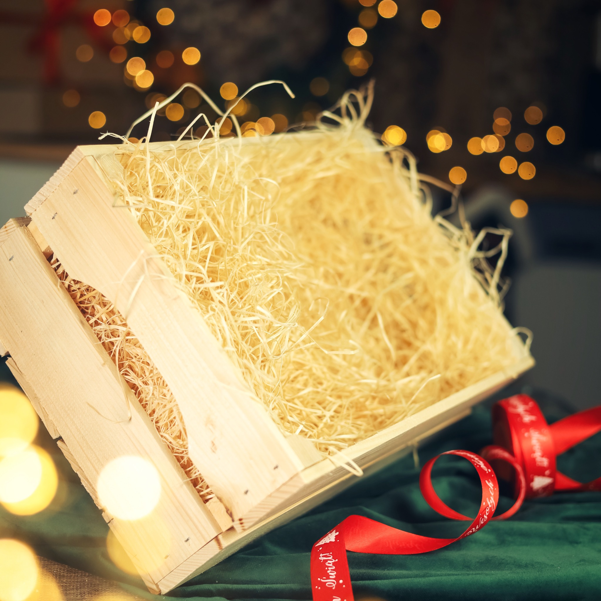 Pusta, jasna, drewniana skrzynka wypełniona jasnym wypełniaczem, obok czerwona wstążka z napisem 'Wesołych Świąt', na zielonym tle z rozmytymi światełkami.