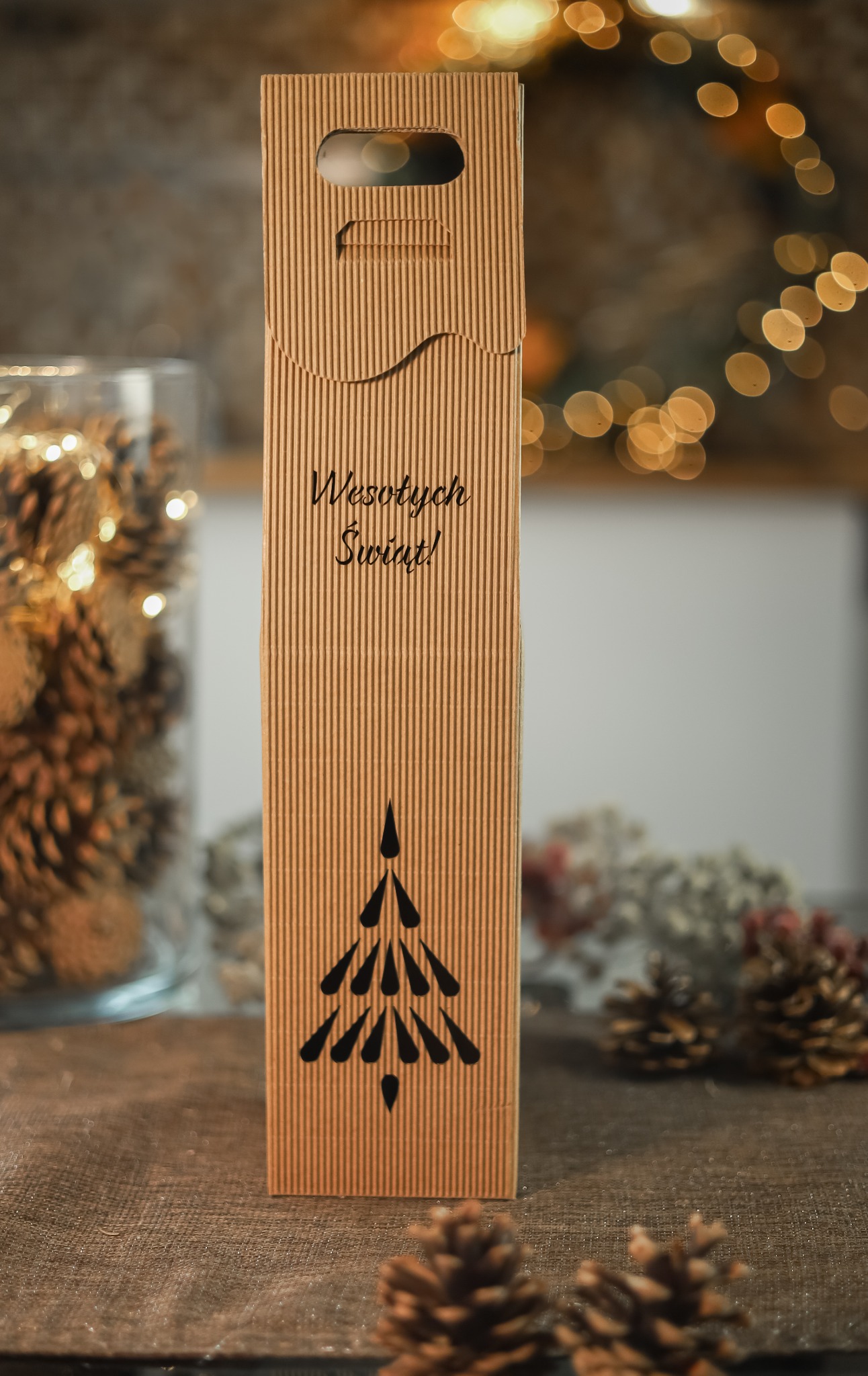 Kartka z życzeniami 'Wesołych Świąt!' na tekturowym opakowaniu prezentowym z motywem choinki, otoczona szyszkami i dekoracyjnymi światełkami w tle, prezentowana w Warszawie.