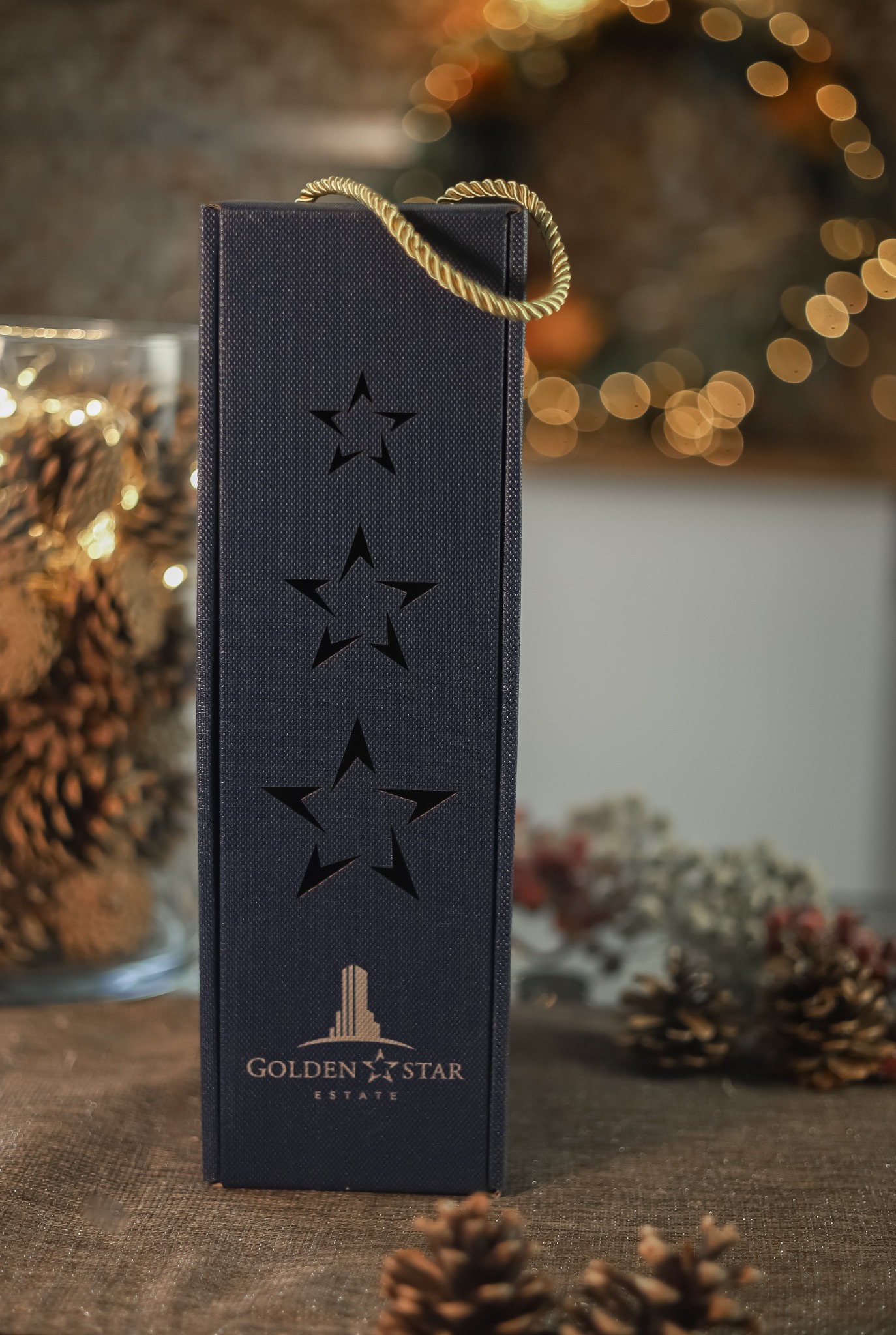 Eleganckie, granatowe pudełko prezentowe na butelkę z wyciętymi gwiazdami i złotym sznurkiem, z logo 'Golden Star Estate' w dolnej części, ustawione na tle z szyszkami i dekoracyjnymi światełkami.