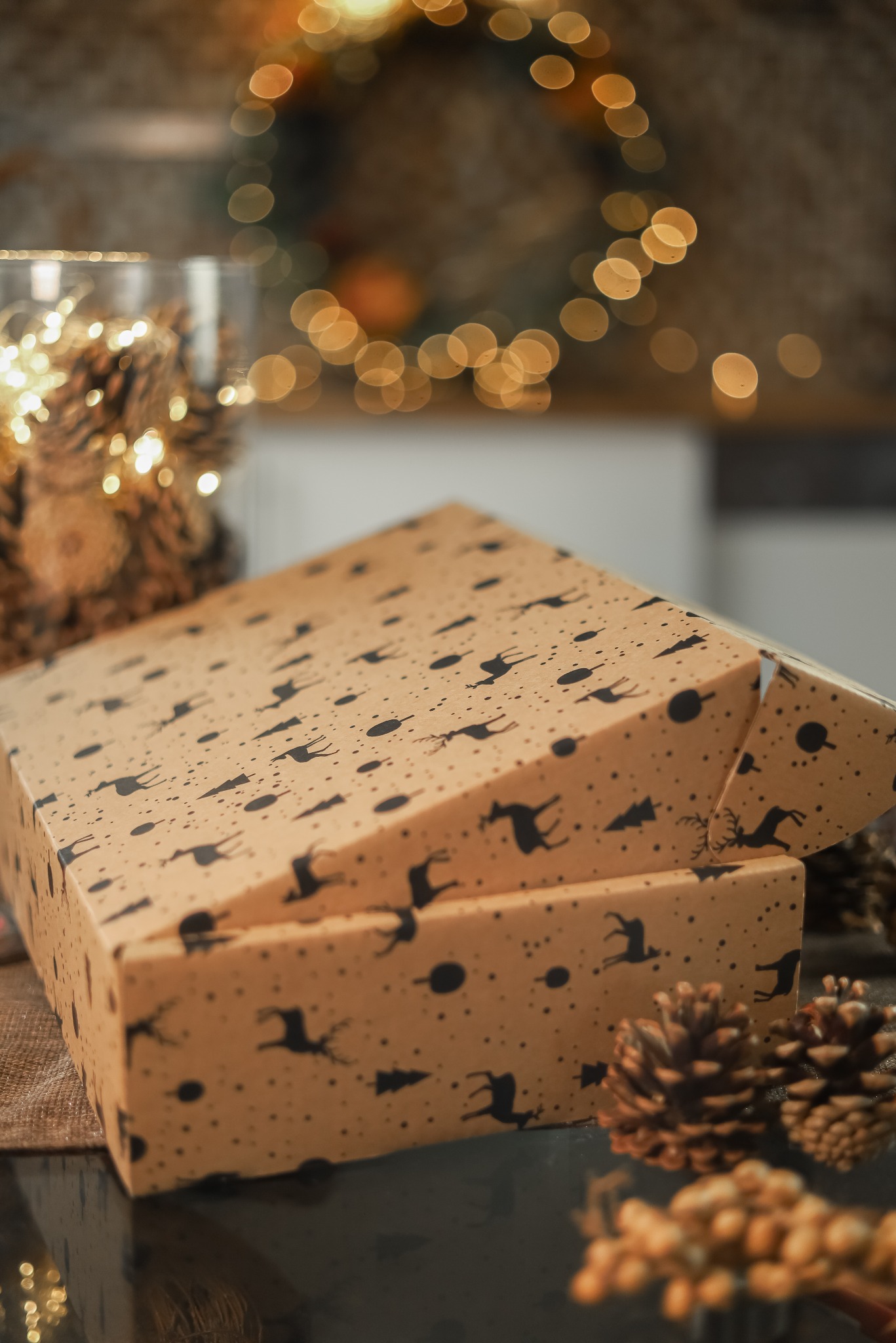 Otwarty prezent w brązowym papierze z czarnymi sylwetkami reniferów i choinek, otoczony szyszkami i delikatnymi światełkami, z wieńcem z lampek w tle.