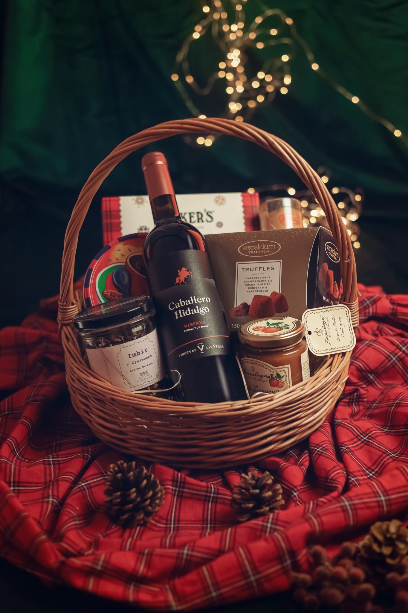 Wigilijny kosz prezentowy z winem Caballero Hidalgo, truflami Excelcium, dżemami Beirobaża i herbatą Imbir z Cynamonem, umieszczony na czerwonej kraciastej tkaninie z szyszkami i światełkami w tle.