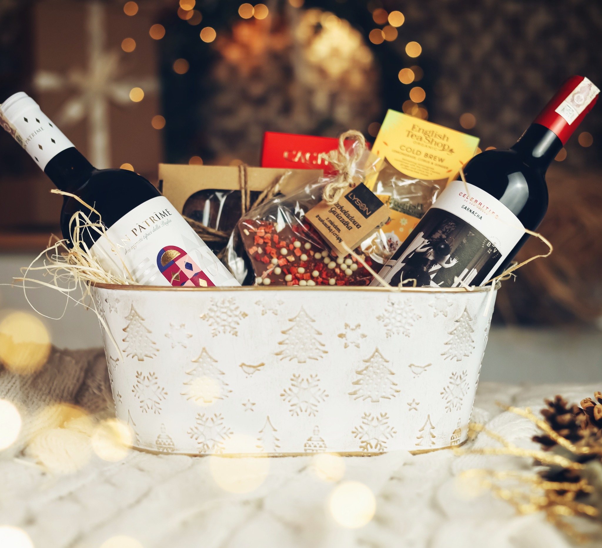 Dekoracyjny, biały kosz z wytłaczanymi motywami choinek i śnieżynek, wypełniony dwiema butelkami czerwonego wina, herbatą English Tea Shop Cold Brew i czekoladową gwiazdką Lyson z miodem...