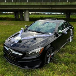 PR1M0 Damian Cybulski - Czarny Mercedes CLA udekorowany białymi kwiatami i tiulem, stojący na trawie pod wiaduktem.