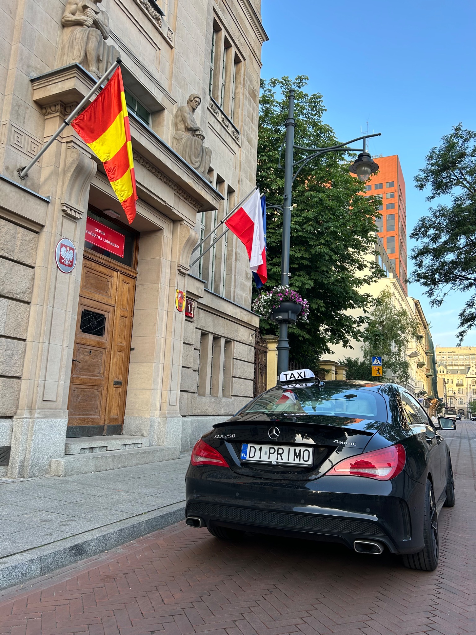 Czarny Mercedes CLA 250 Taxi z tablicą rejestracyjną D1 PRIMO zaparkowany na ulicy przed budynkiem z flagami Hiszpanii i Francji.