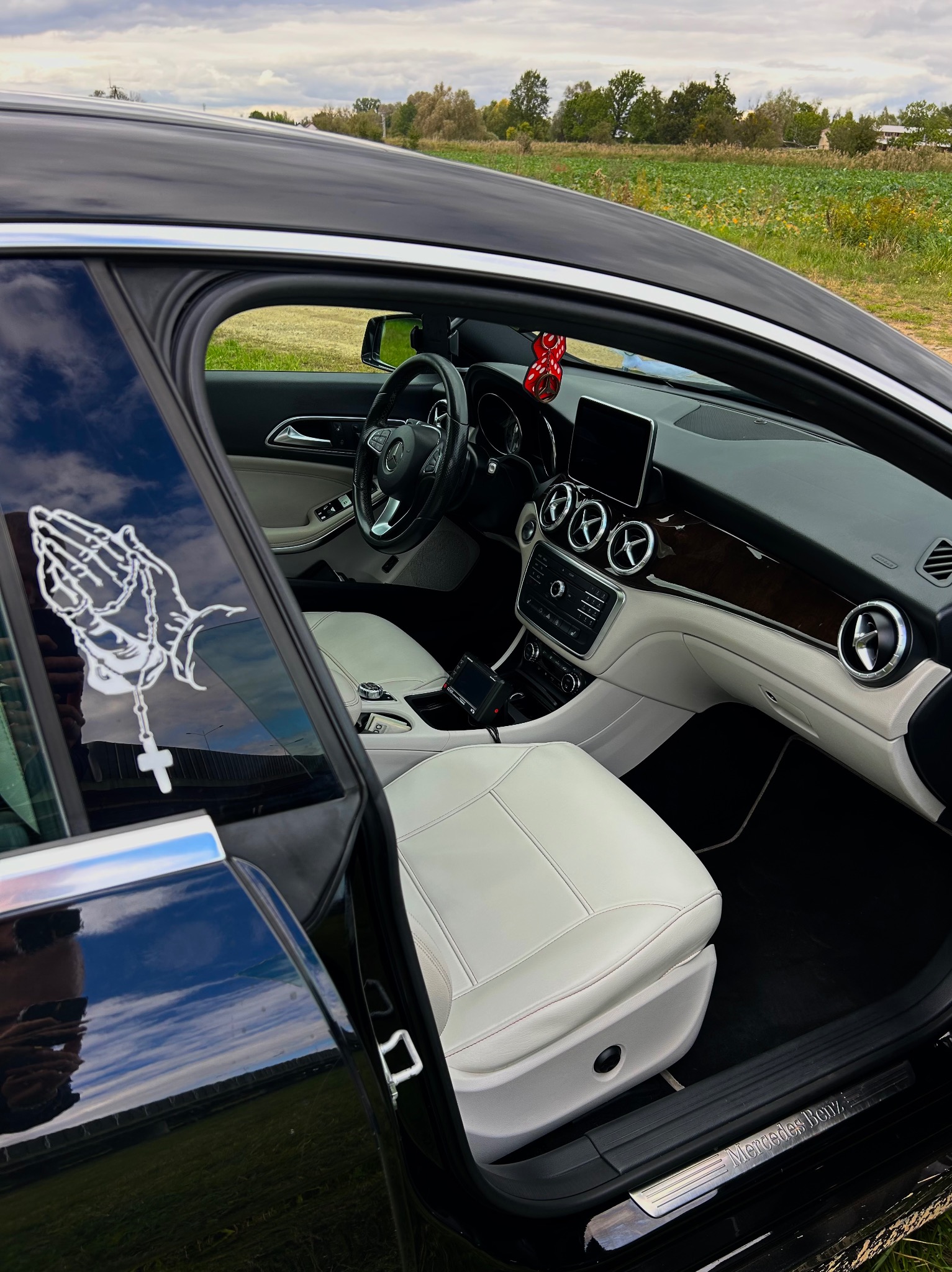 Wnętrze czarnego Mercedesa z jasną skórzaną tapicerką, widoczne przez otwarte drzwi; na szybie naklejka z motywem dłoni w modlitwie z różańcem.