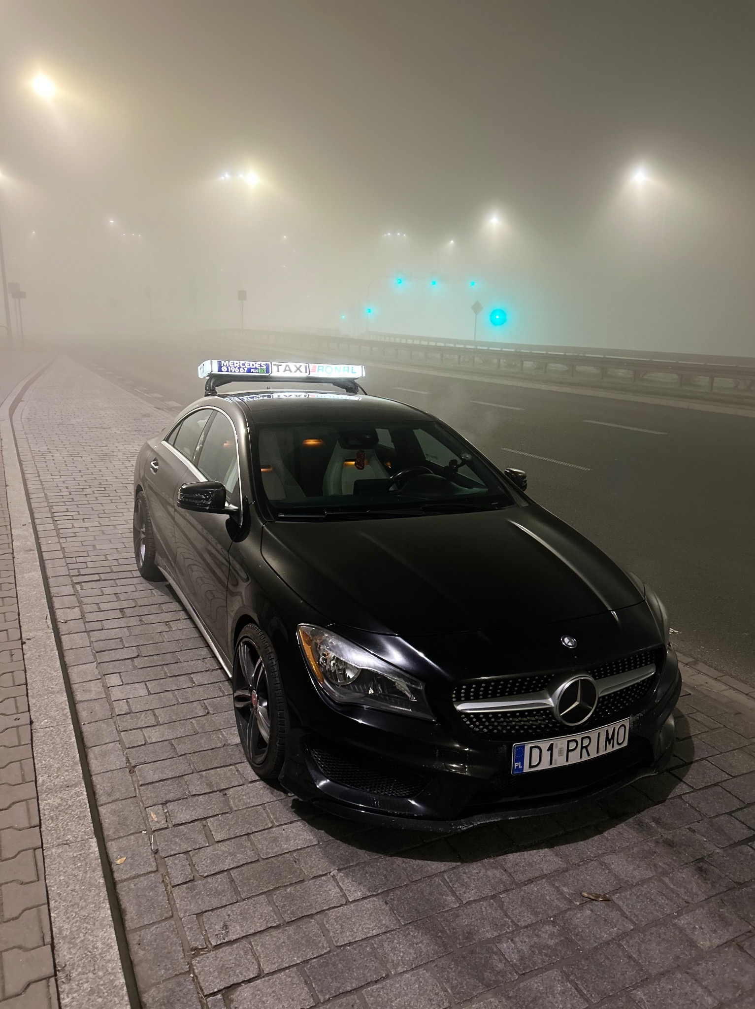 Czarny Mercedes CLA z podświetlanym napisem TAXI RONAL na dachu, zaparkowany na chodniku z kostki brukowej we mgle, widoczne światła uliczne w tle.