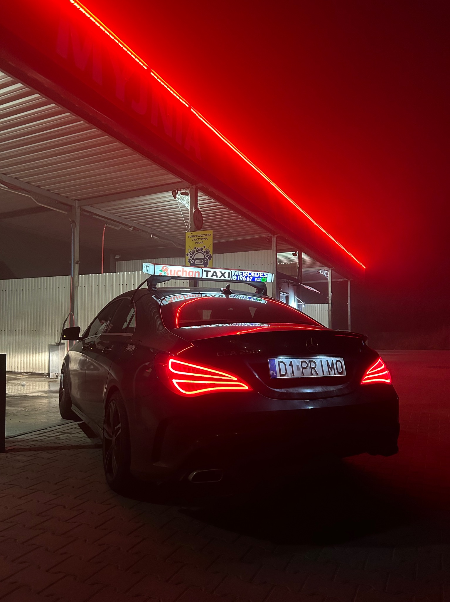 Czarny Mercedes CLA 250 z bagażnikiem dachowym i tabliczką Taxi, zaparkowany pod zadaszeniem z czerwonym oświetleniem neonowym.