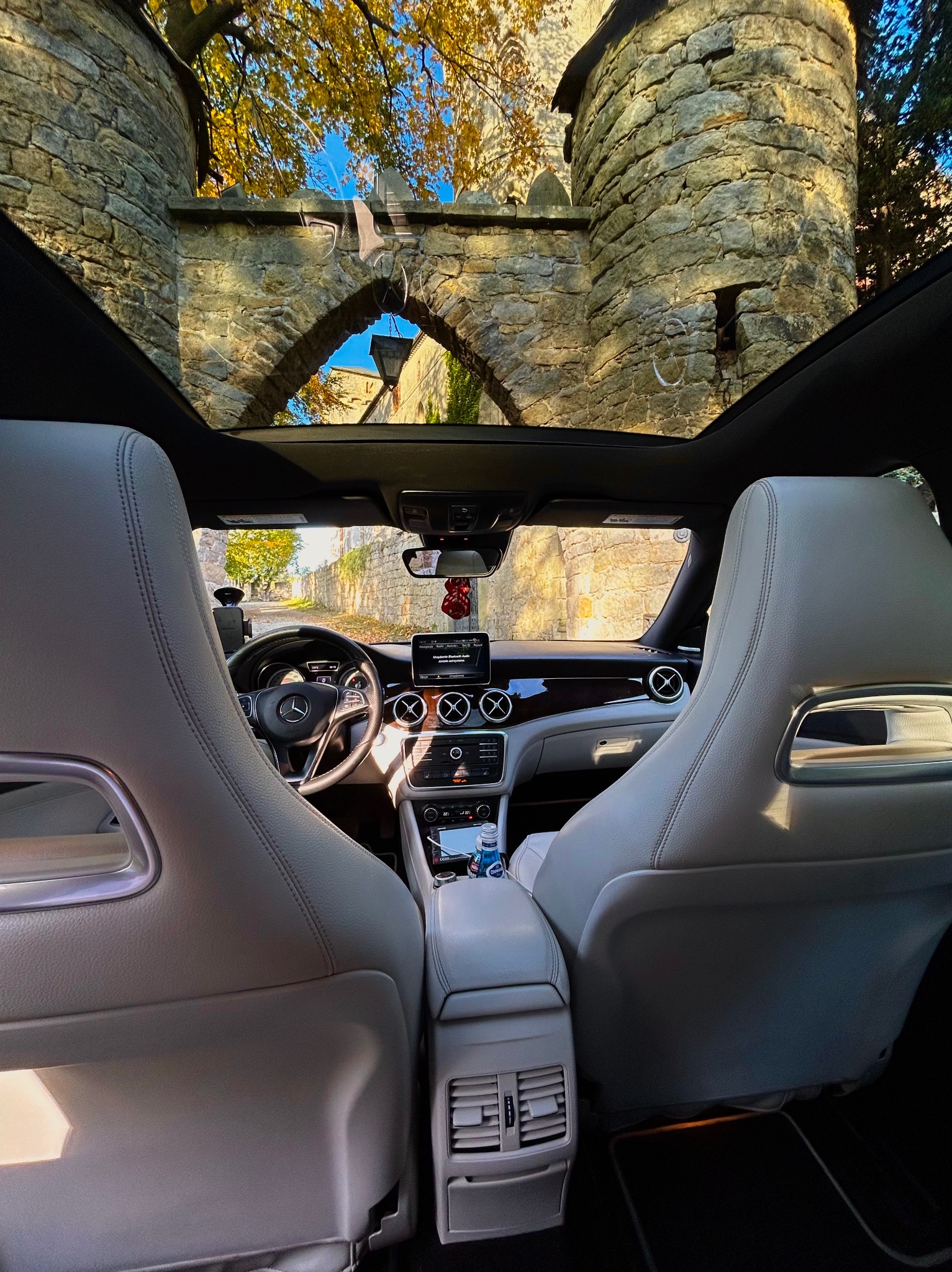 Wnętrze samochodu z widokiem przez panoramiczny dach na średniowieczną kamienną bramę i jesienne drzewa.