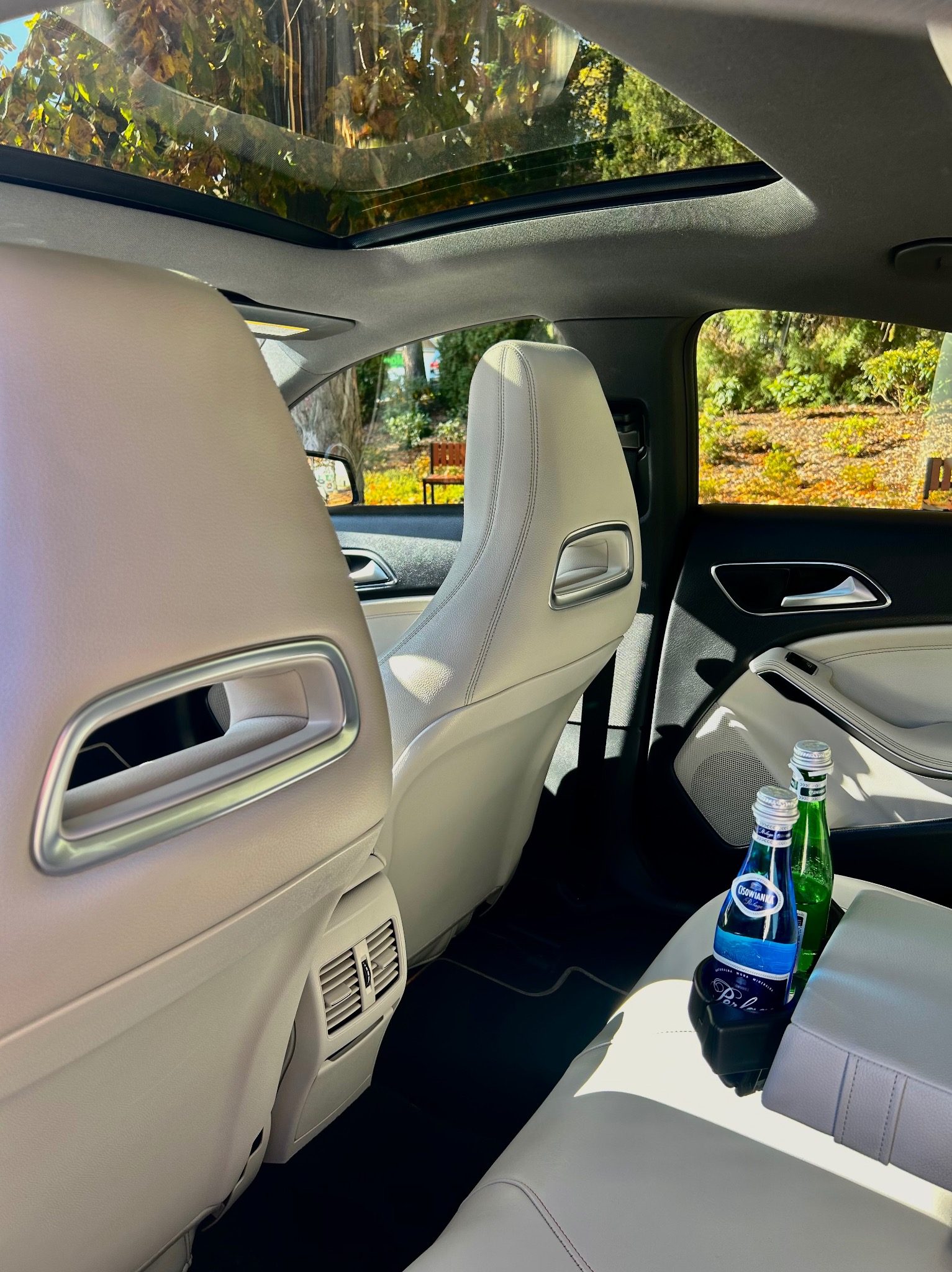 Wnętrze luksusowego samochodu z jasną skórzaną tapicerką, widok z tyłu, panoramiczny dach z widokiem na drzewa, butelki wody w uchwycie.
