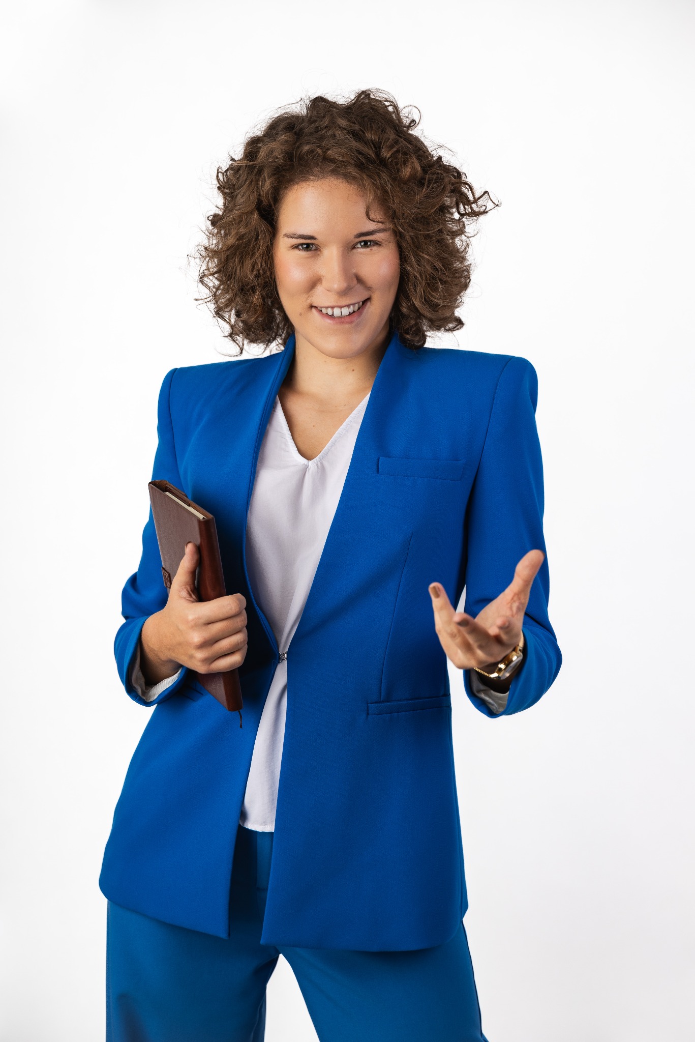 Uśmiechnięta kobieta w niebieskim garniturze prezentuje otwartą dłonią, trzymając brązowy notatnik.