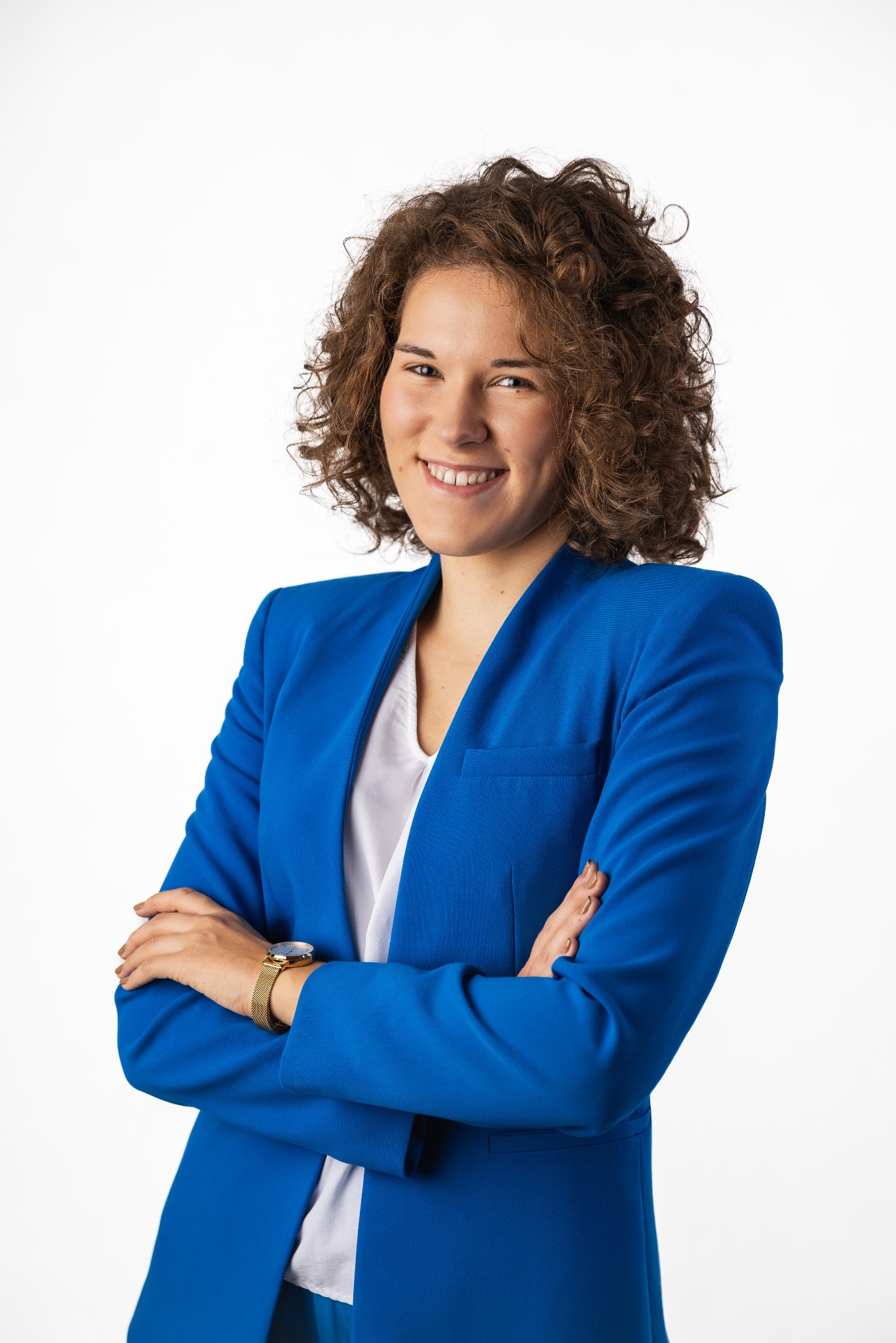 Uśmiechnięta kobieta z kręconymi włosami, ubrana w niebieski żakiet i białą bluzkę, pozuje z założonymi rękami na białym tle. Na nadgarstku widoczny złoty zegarek.