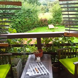 Pieśń Duszy- holistyczne wsparcie dla ciała, ducha i umysłu. - Relaksujący widok z balkonu z zielonymi poduszkami na krzesłach, wiszącymi dzwonkami wietrznymi i widokiem na zielony ogród w tle.