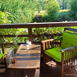 Pieśń Duszy- holistyczne wsparcie dla ciała, ducha i umysłu. - Drewniany taras z meblami ogrodowymi: stół z dekoracjami, krzesła z jaskrawozielonymi poduszkami, roślina w doniczce, widok na zieleń za balustradą.