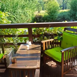 Drewniany taras z meblami ogrodowymi: stół z dekoracjami, krzesła z jaskrawozielonymi poduszkami, roślina w doniczce, widok na zieleń za balustradą.