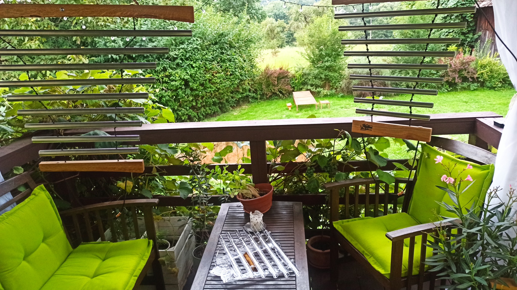 Relaksujący widok z balkonu z zielonymi poduszkami na krzesłach, wiszącymi dzwonkami wietrznymi i widokiem na zielony ogród w tle.