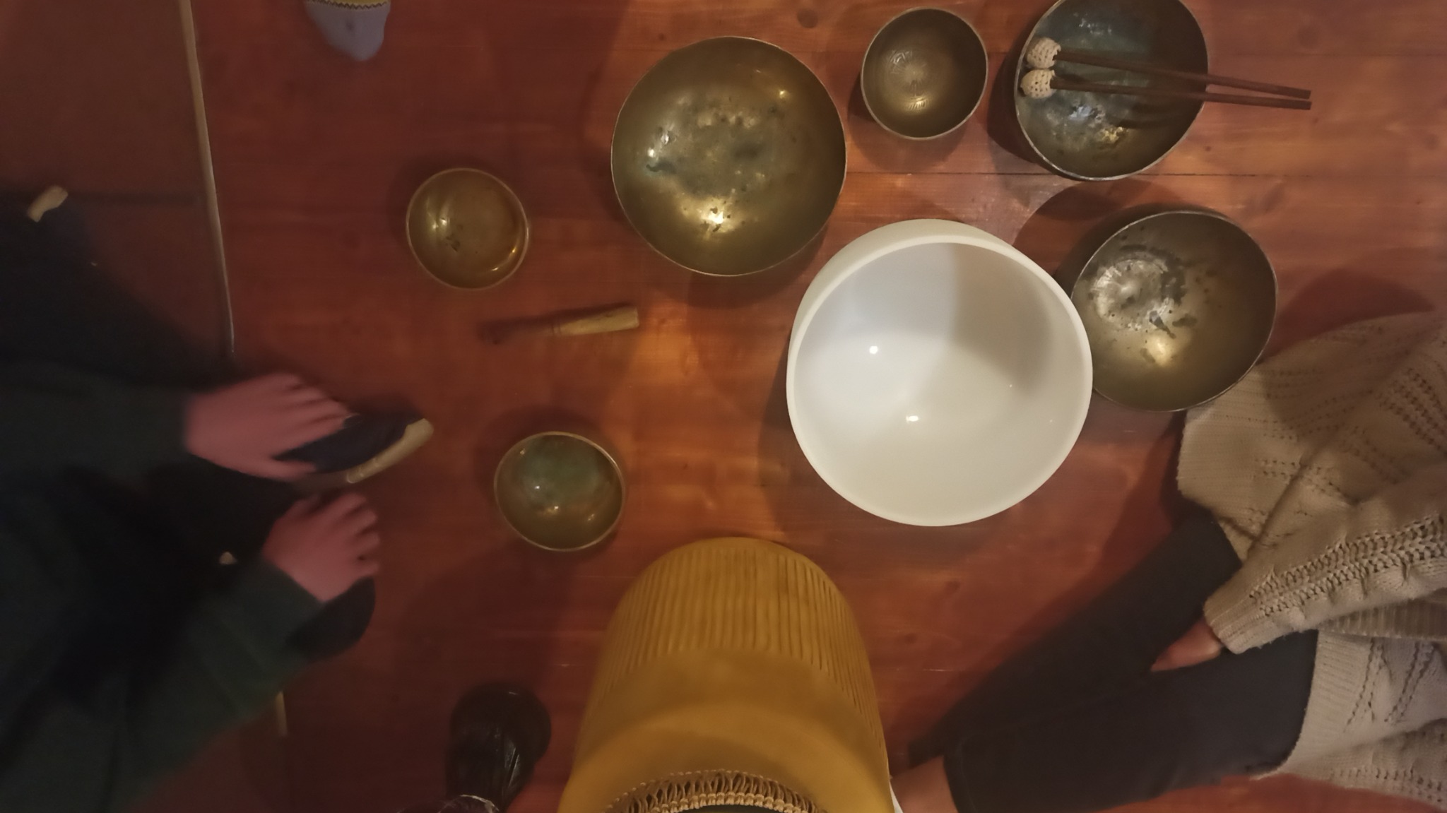 Różne misy dźwiękowe, w tym jedna duża biała, rozłożone na drewnianym stole, z widocznymi dłońmi osób przygotowujących się do sesji relaksacyjnej.