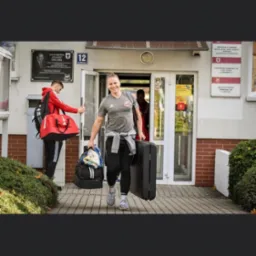 Uśmiechnięty mężczyzna z blond irokezem, w sportowym stroju, wychodzi z budynku niosąc walizkę i torbę sportową; drugi mężczyzna w czerwonej bluzie z torbą wchodzi do budynku.