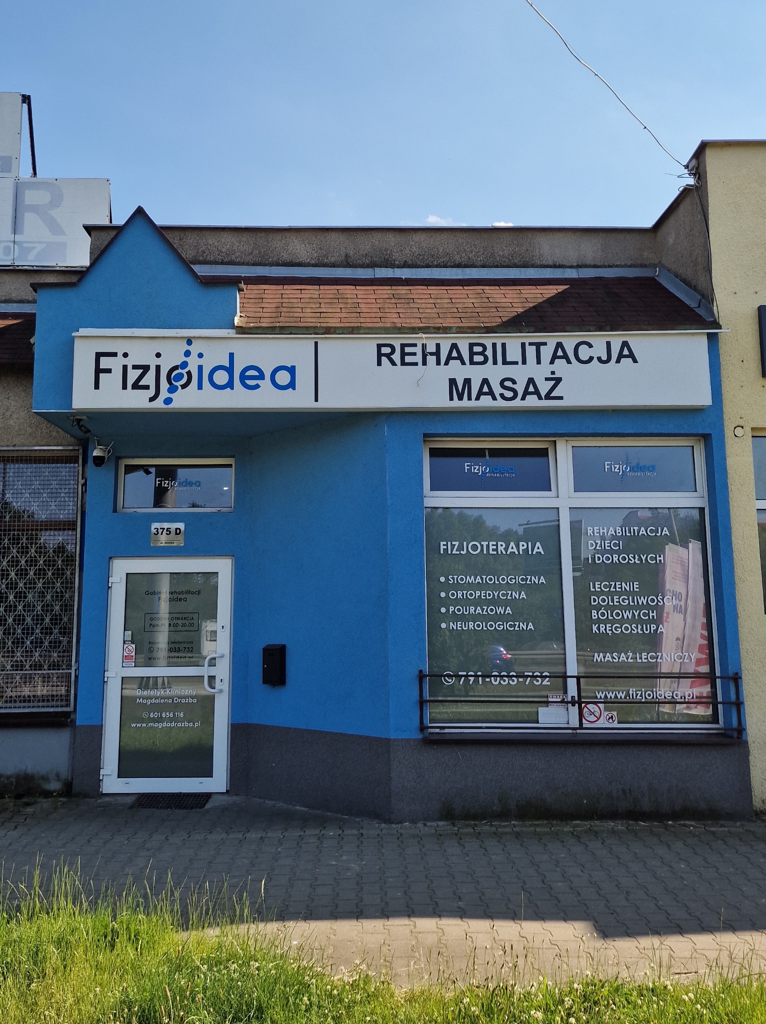 Fasada budynku w kolorze niebieskim z szyldem 'Fizjoidea | Rehabilitacja Masaż'. W oknach plakaty informujące o fizjoterapii i rehabilitacji dzieci i dorosłych. Widoczne godziny otwarcia i numer...