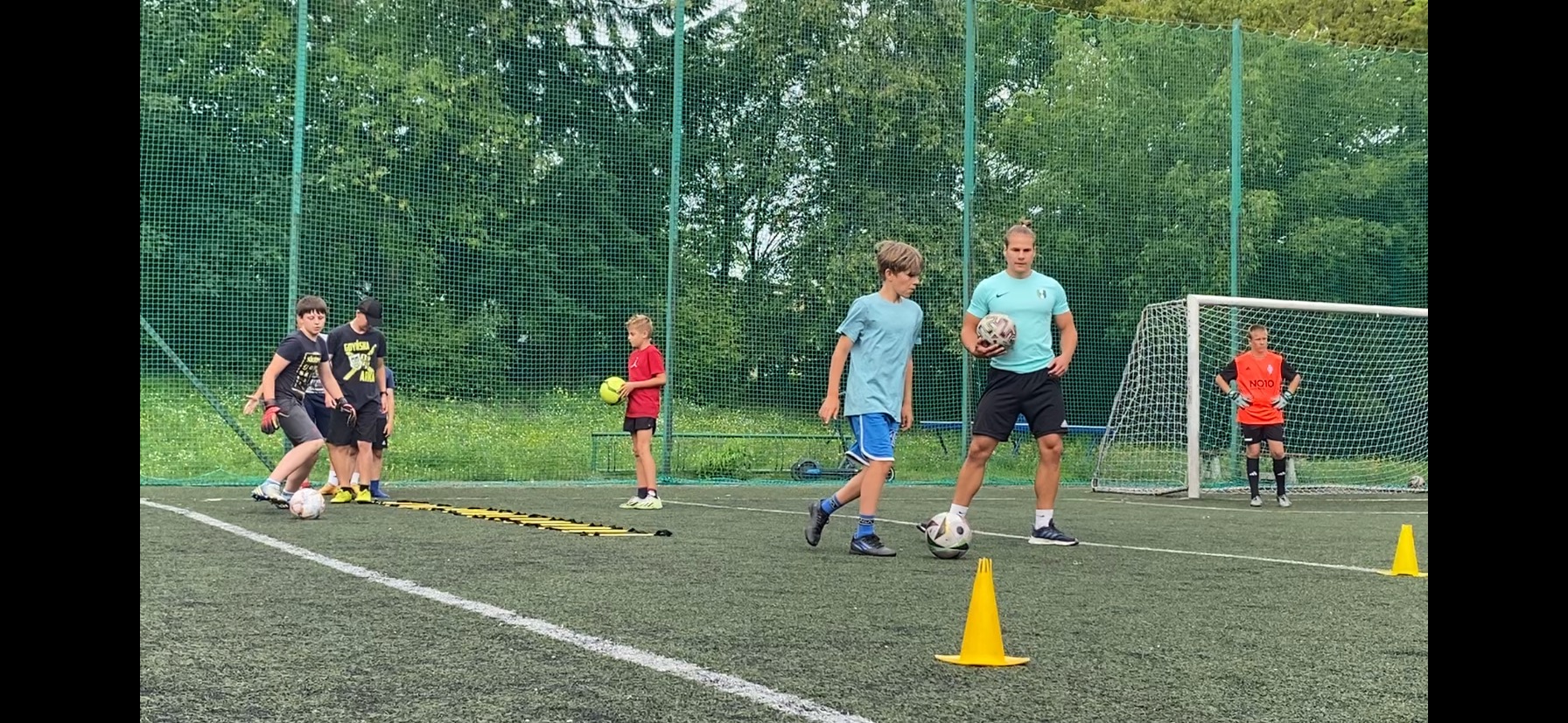 Trening piłkarski na zielonym boisku: grupa chłopców ćwiczy pod okiem trenera, widoczne bramka, piłki i żółte pachołki.