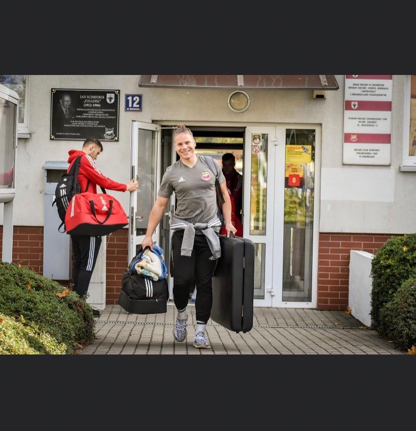 Uśmiechnięty mężczyzna z blond irokezem, w sportowym stroju, wychodzi z budynku niosąc walizkę i torbę sportową; drugi mężczyzna w czerwonej bluzie z torbą wchodzi do budynku.