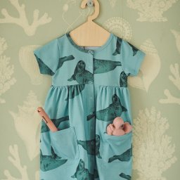 AIDY - Produkcja Odzieży - Błękitna sukienka dziecięca z motywem fok, wisząca na drewnianym wieszaku na tle ściany w morskie wzory. W kieszeniach sukienki widać dziecięce zabawki.