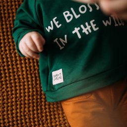 AIDY - Produkcja Odzieży - Niemowlę leżące na brązowym kocu w zielonej bluzie z napisem 'We blossom in the wild' i metką 'Born to explore', ubrane w pomarańczowe spodnie.