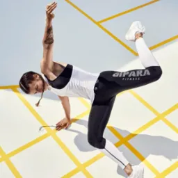 Kobieta w sportowym stroju (czarne legginsy z białym napisem, biała koszulka) w dynamicznej pozie na białym tle z żółtymi liniami, widoczny tatuaż na ramieniu.