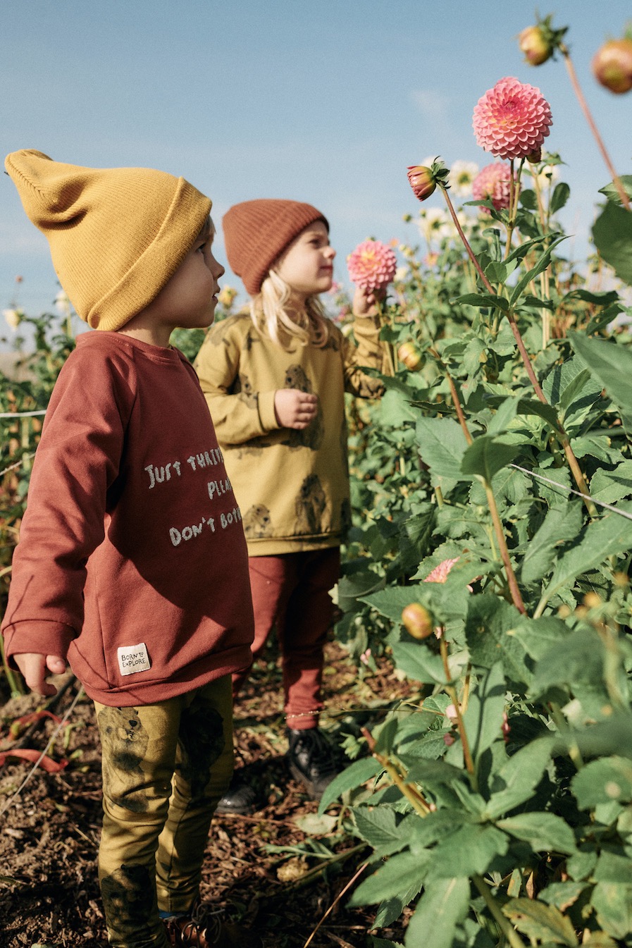 Dwoje dzieci w jesiennych ubraniach stoi wśród kwitnących dalii; chłopiec w bordowej bluzie z napisem 'Just Thriving Please Don't Bother' i dziewczynka w musztardowej bluzie z motywem lwa.