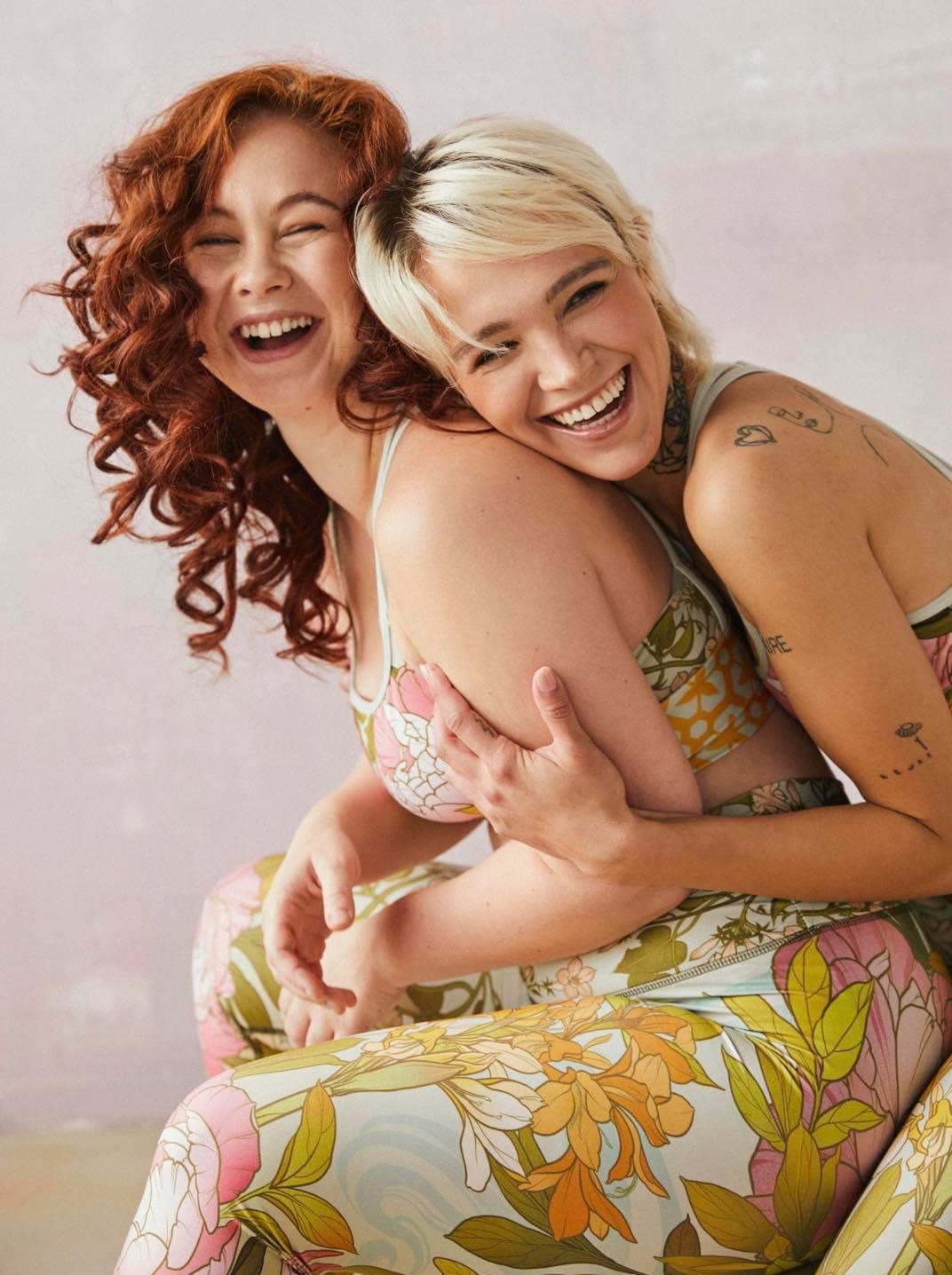 Dwie uśmiechnięte kobiety, jedna z rudymi kręconymi włosami, druga z blond krótkimi, obejmują się, ubrane w sportowe topy i legginsy w kwiatowe wzory na tle delikatnego różowego tła.