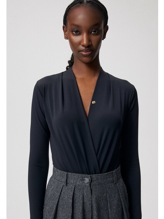 Kobieta pozuje w czarnym body z długim rękawem i drapowaniem na dekolcie, uzupełnionym złotą broszką, oraz w szarej spódnicy w drobną kratę.