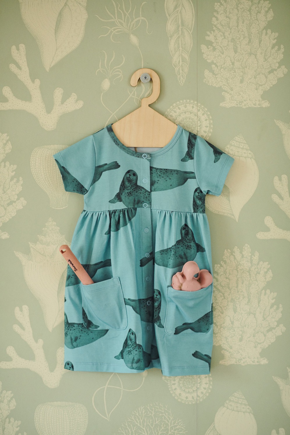 Błękitna sukienka dziecięca z motywem fok, wisząca na drewnianym wieszaku na tle ściany w morskie wzory. W kieszeniach sukienki widać dziecięce zabawki.