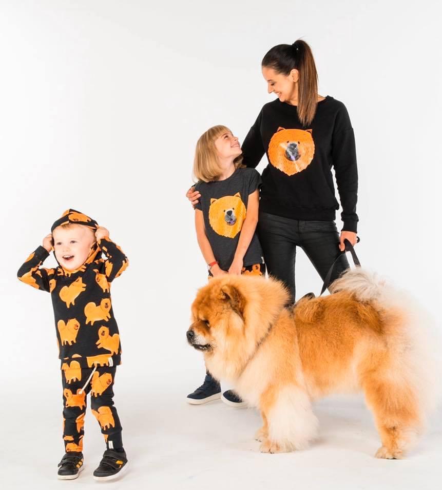 Rodzina w pasujących strojach z nadrukiem Chow Chow, dziecko w dresie z kapturem, dziewczynka w koszulce, mama w bluzie, obok nich pies rasy Chow Chow, białe tło.