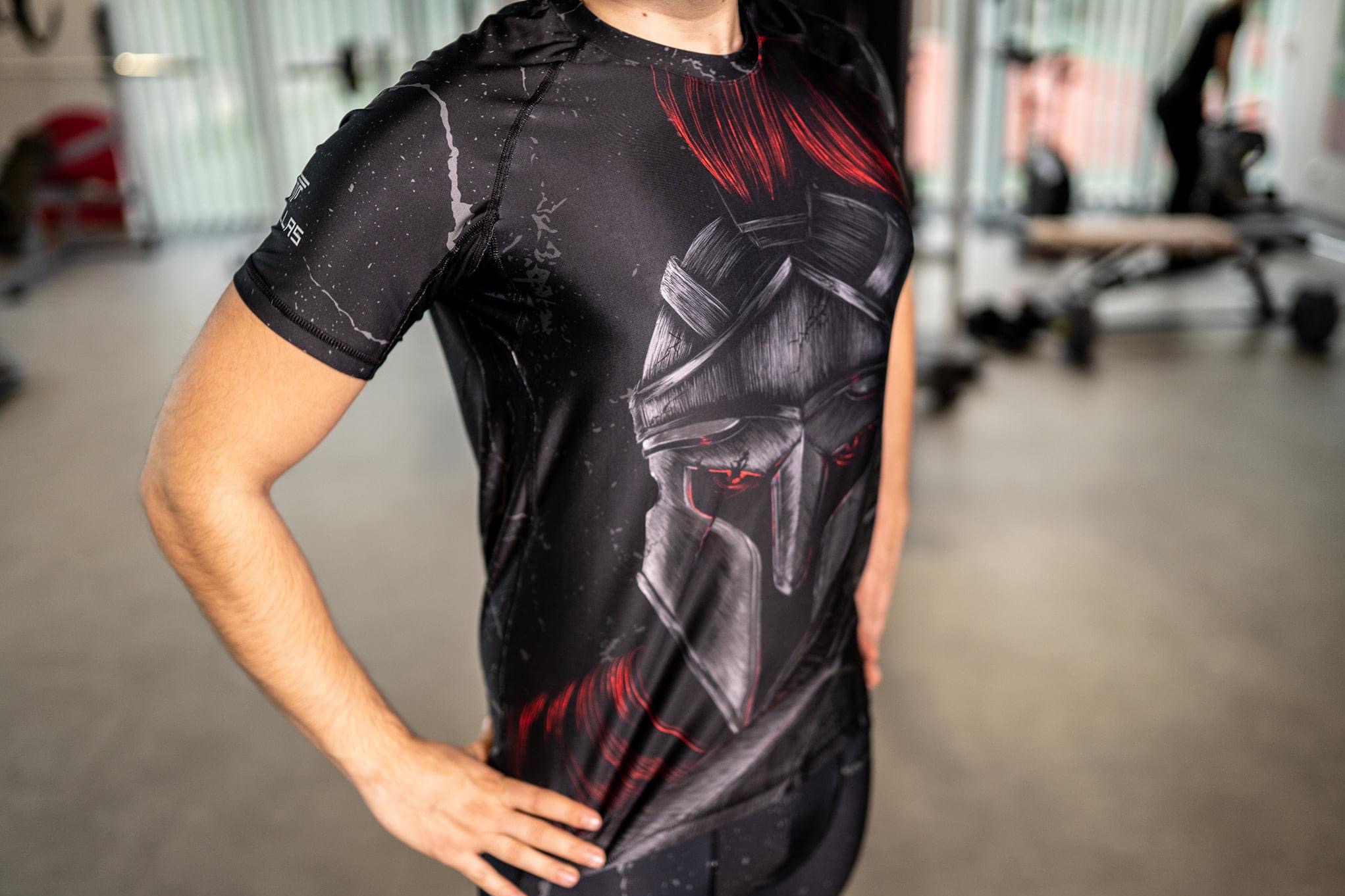 Mężczyzna w czarnej koszulce termoaktywnej z nadrukiem hełmu spartańskiego w odcieniach szarości i czerwieni, pozujący w siłowni.