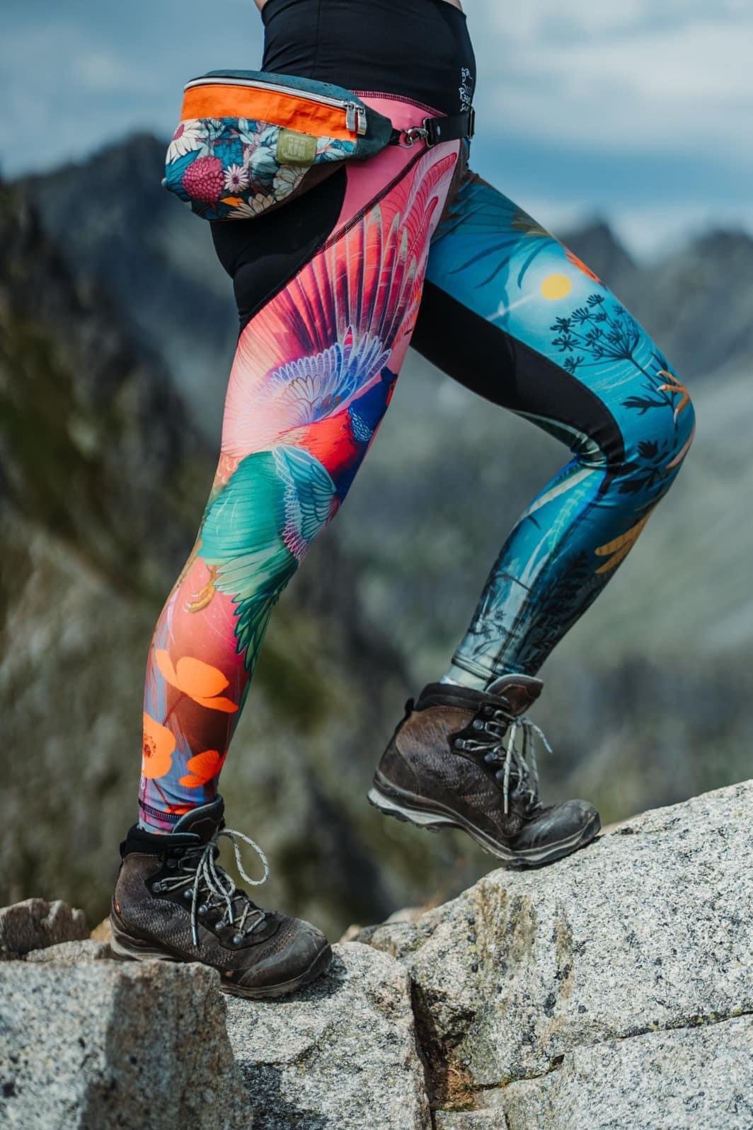 Osoba w górach, ubrana w legginsy z nadrukiem ptaków i roślin, z saszetką biodrową w kwiaty, w trekkingowych butach na kamieniu.