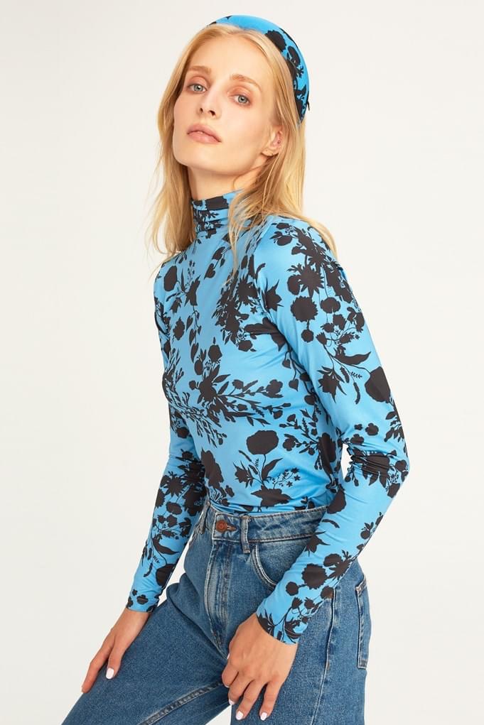 Modelka w błękitnej bluzce z czarnym kwiatowym wzorem i dżinsach, pozująca na jasnym tle.